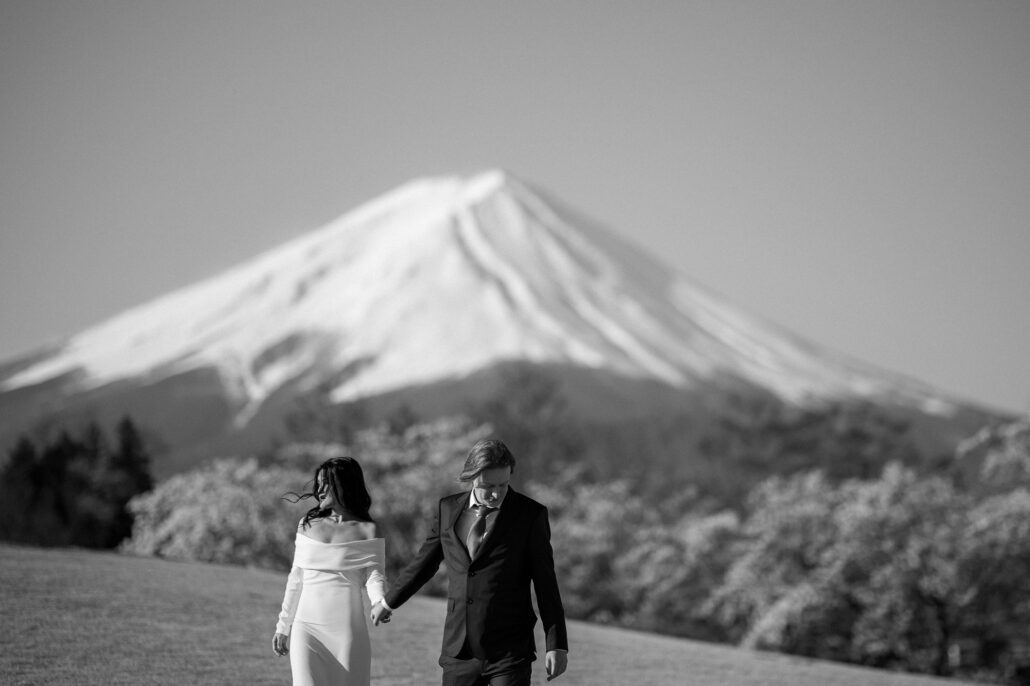 In black and white, the couple walk hand in hand across open grass below snow-capped Mt Fuji.