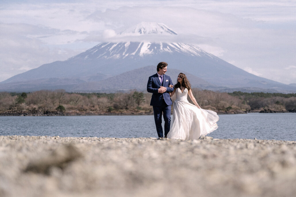 The bride and groom walk along the stony shore of Lake Shoji, her skirt swinging, beneath a snow-capped Mt Fuji on their elopement day.