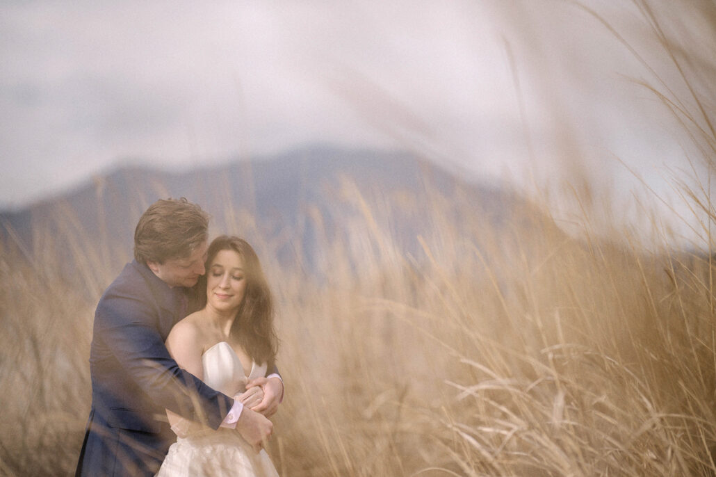 The groom holds the bride from behind as she smiles in tall golden grass, misty mountains behind, a Mt Fuji elopement portrait.