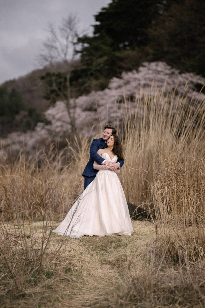 The groom wraps his arms around the bride in tall golden grass beside a blossoming cherry tree, a spring elopement near Mt Fuji.