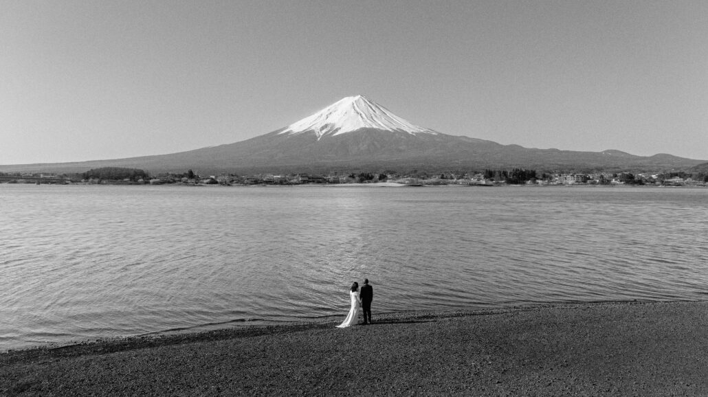In black and white, a couple stand small at the water's edge across the lake from Mt Fuji.
