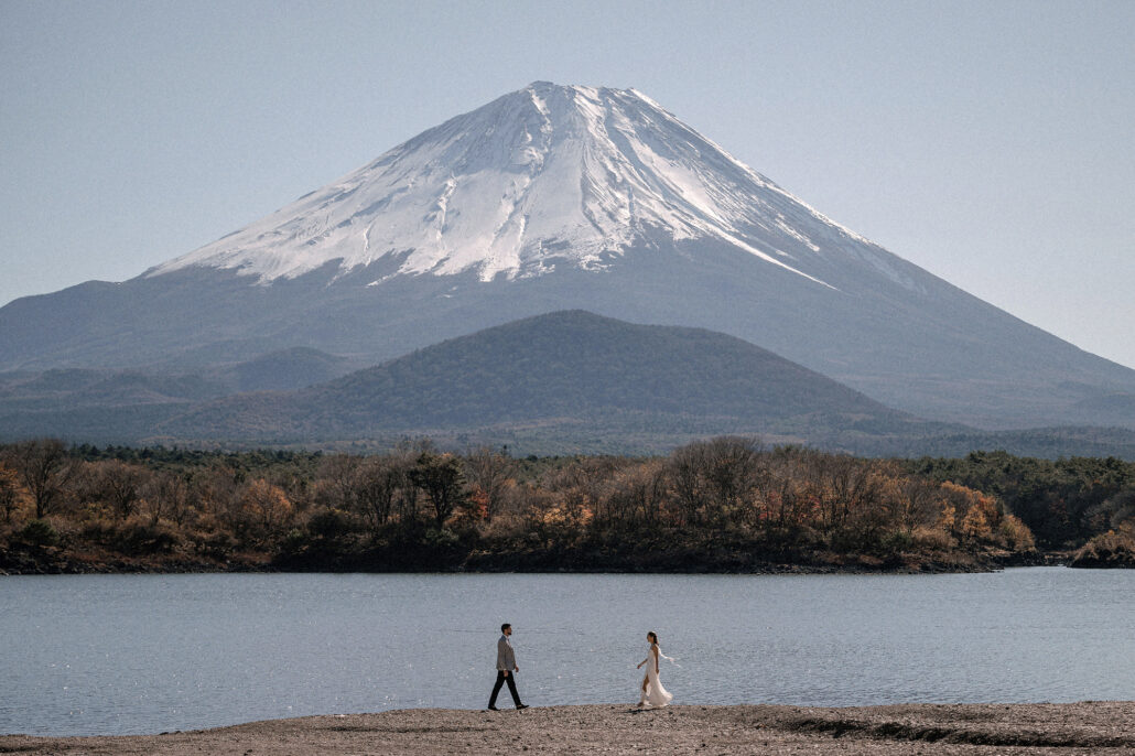 The bride and groom walk along a lakeshore beneath a towering, snow-capped Mt Fuji in autumn.