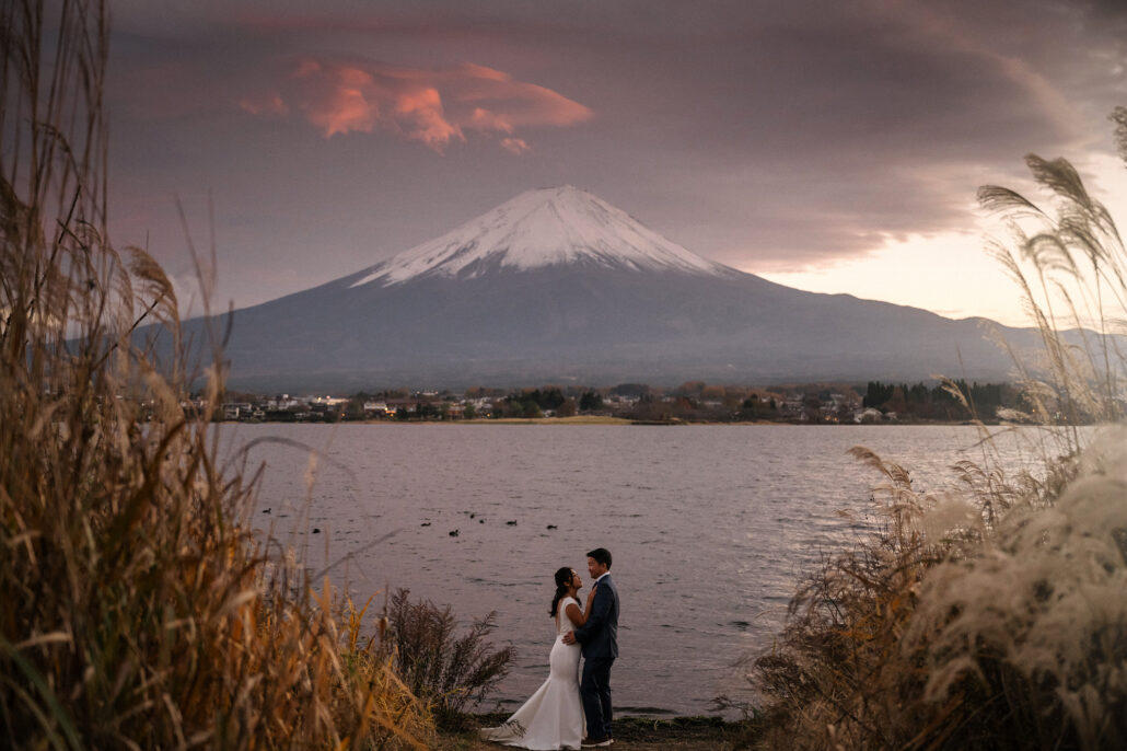 Framed by silver grass, a bride and groom hold each other on a lakeshore below Mt Fuji as pink cloud drifts over the summit.