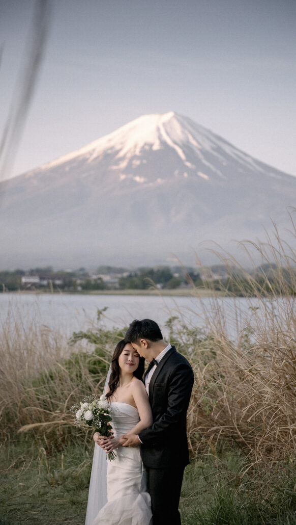At sunrise a couple embrace among the grasses on the shore of Lake Kawaguchi, Mt Fuji clear and snow-capped across the water.