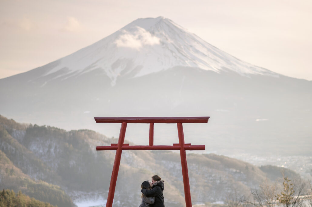 A couple in winter coats kiss beneath a red torii on a hillside, snow-capped Mt Fuji rising behind them through a soft haze.