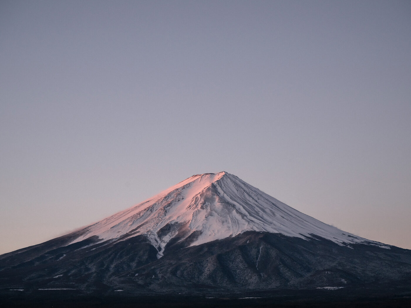 The snowcap of Mt Fuji glows pink in the first light of a winter morning, the sky still pale above it.
