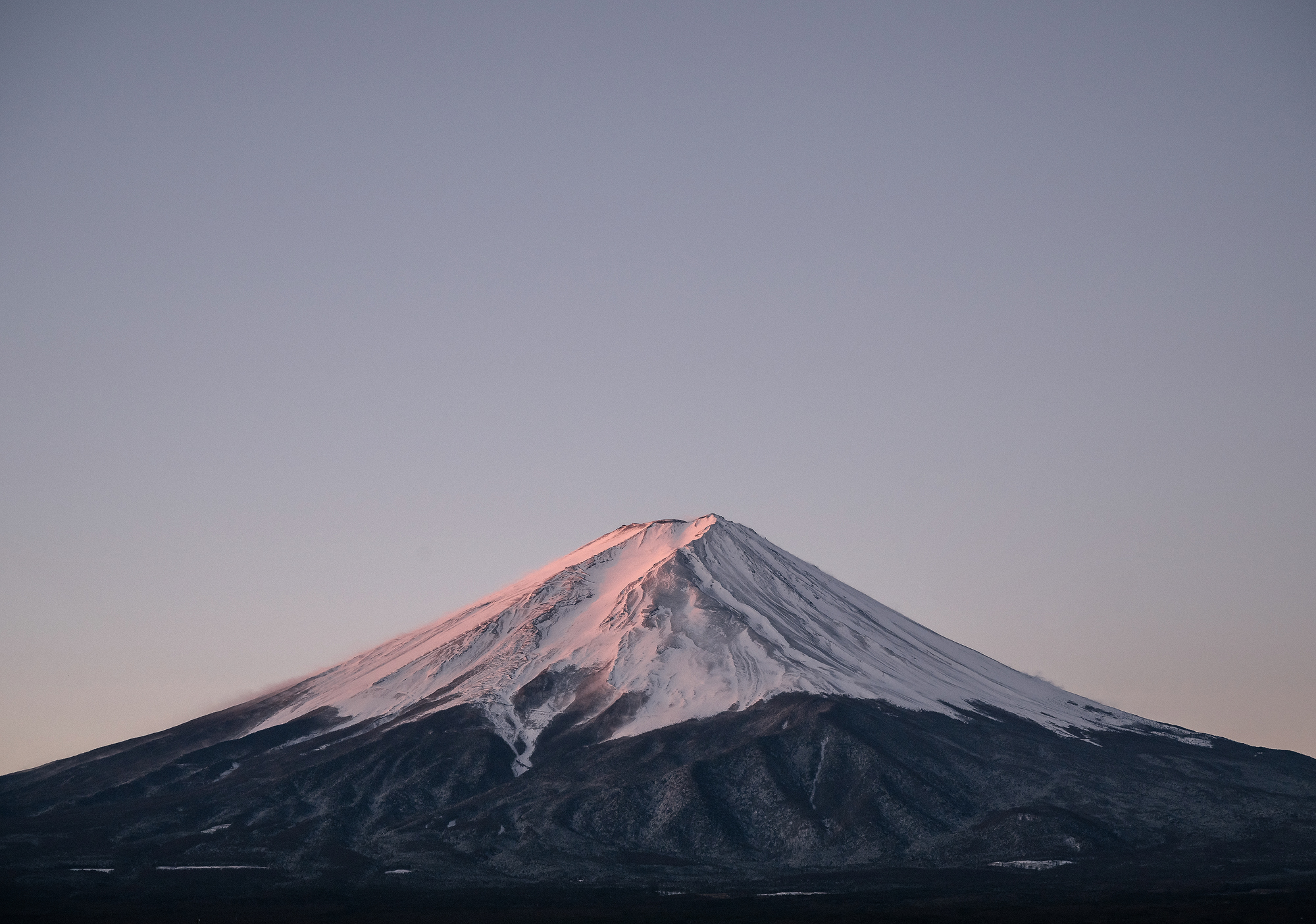 The snowcap of Mt Fuji glows pink in the first light of a winter morning, the sky still pale above it.