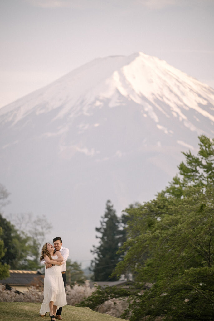 The couple embraces, laughing, on a grassy slope below Mt Fuji, its snowy summit towering over the green spring trees.