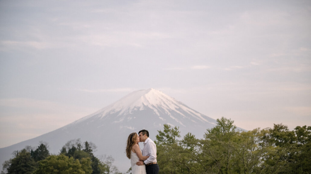 The couple kisses above a line of green treetops, Mt Fuji in spring pale against the soft evening sky at Kawaguchiko.