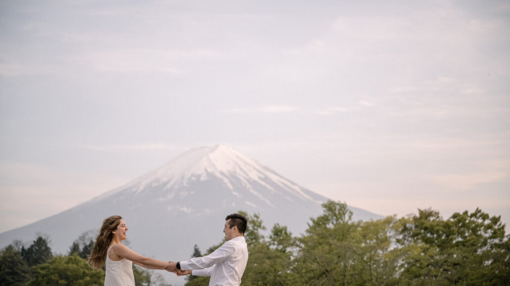 Laughing, the couple swings hands at arm's length above the green trees, Mt Fuji in spring rising behind them at dusk.