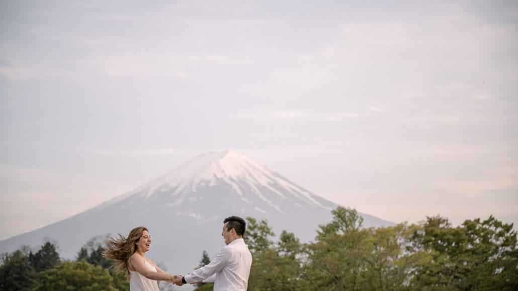 Her hair flying in the wind, the couple holds hands at arm's length above green trees, Mt Fuji white against the evening sky.