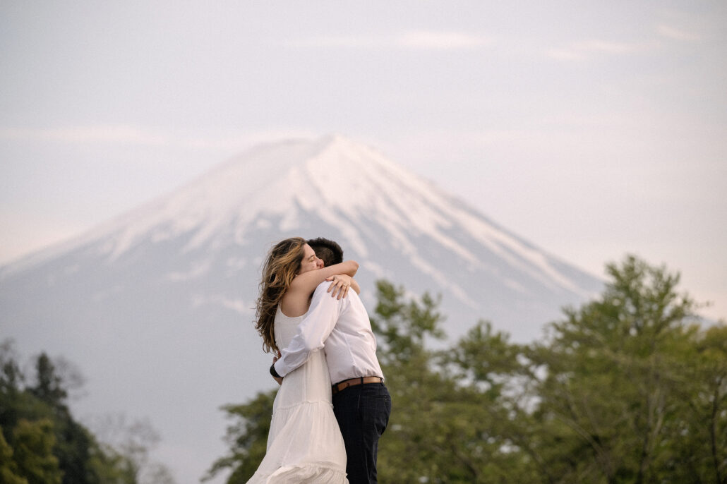 The couple hugs tightly in the evening light, her white dress catching the breeze, Mt Fuji's snowy slopes softly blurred.