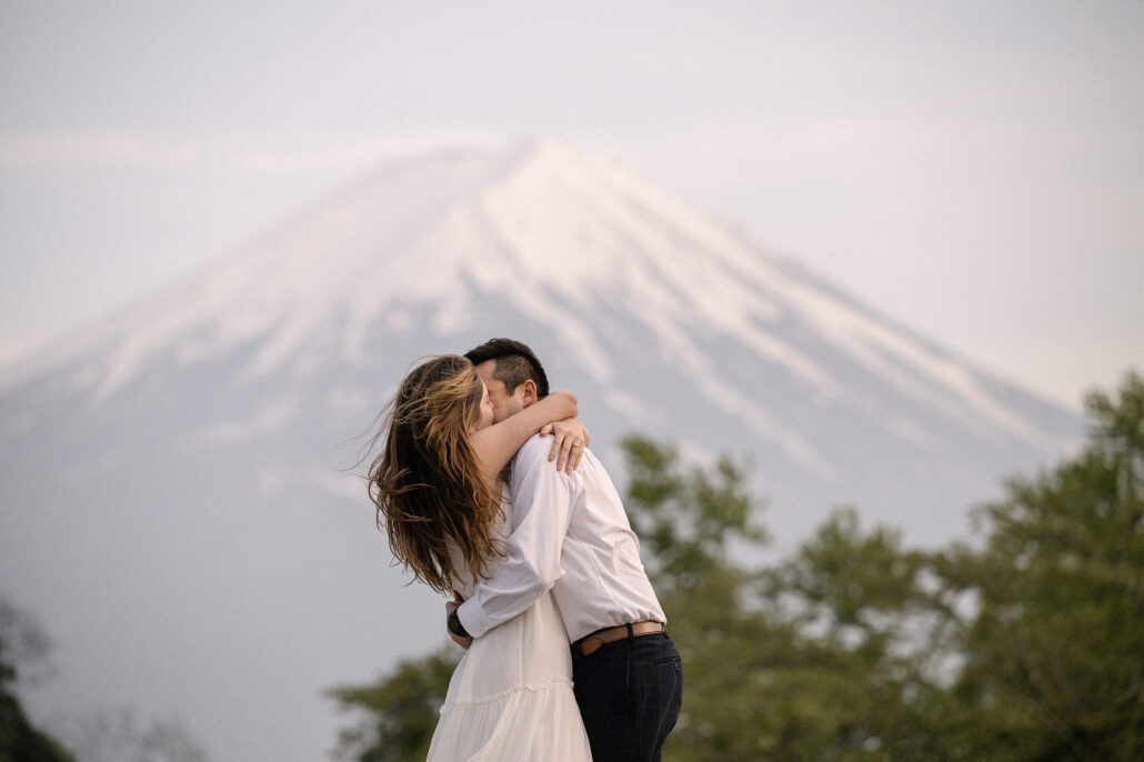 Wind lifts her hair as the couple kisses in the evening, the snow-covered slopes of Mt Fuji soft behind the green trees.