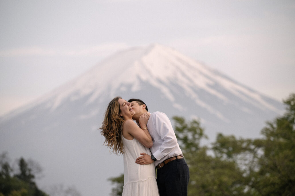 She tips her head back laughing as he holds her close, Mt Fuji soft and snow-covered above the trees after their proposal.