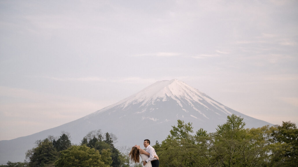 A wide view of the couple laughing as she leans back in his arms above the treetops, Mt Fuji in spring snow under a soft sky.