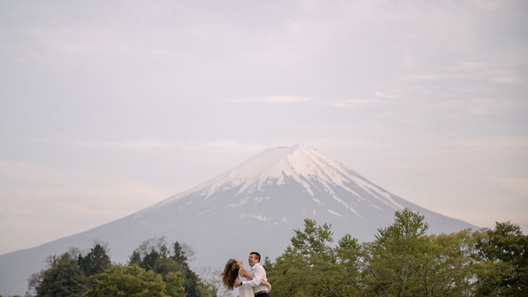 A wide evening view of the couple laughing in an embrace above green spring trees, Mt Fuji snow-capped against a pale sky.