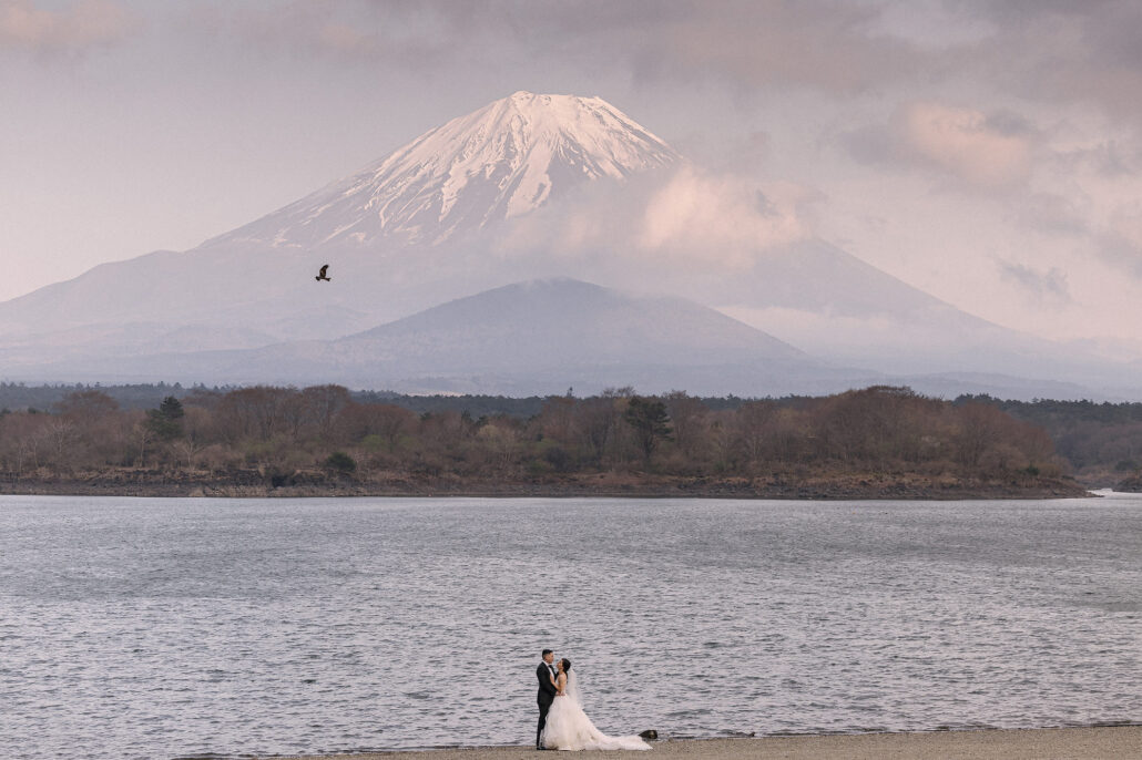 A bride and groom stand alone on a lakeshore at Mt Fuji, small against the water, the summit white with snow behind them.