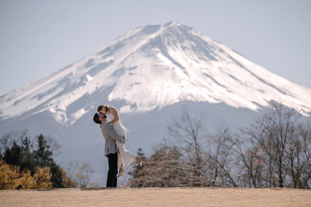 The groom lifts the bride high as they kiss in front of snow-capped Mt Fuji in late autumn.