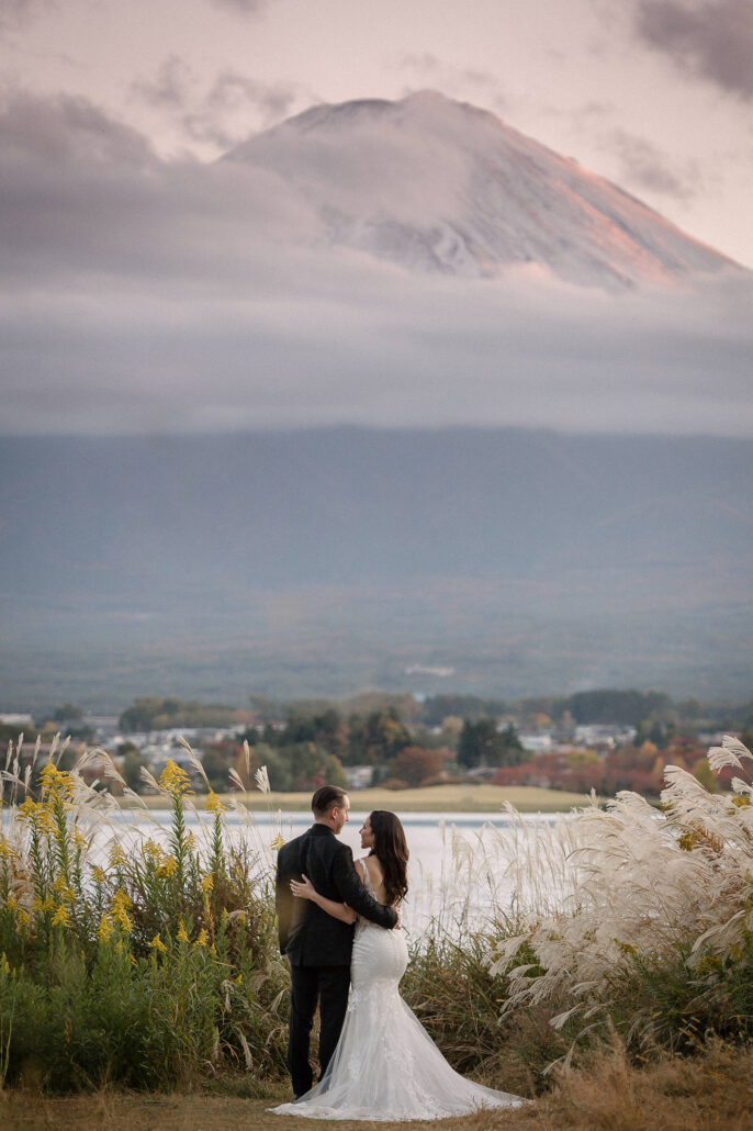 The groom holds the bride on the lakeshore under a pink dusk sky as cloud wraps the snowy summit of Mt Fuji above Lake Kawaguchiko.