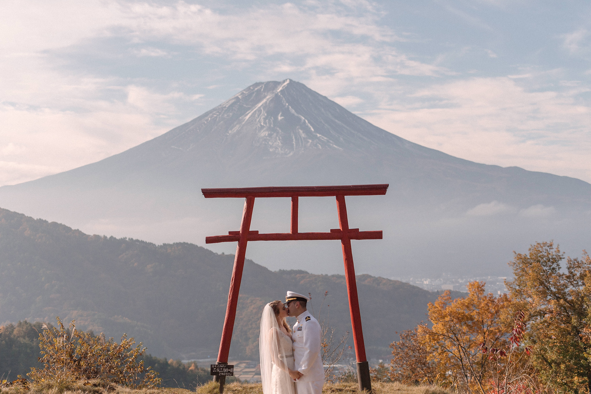 The couple stand small beneath a red torii gate with snow-capped Mt Fuji behind, Mt Fuji wedding portraits on an autumn morning.