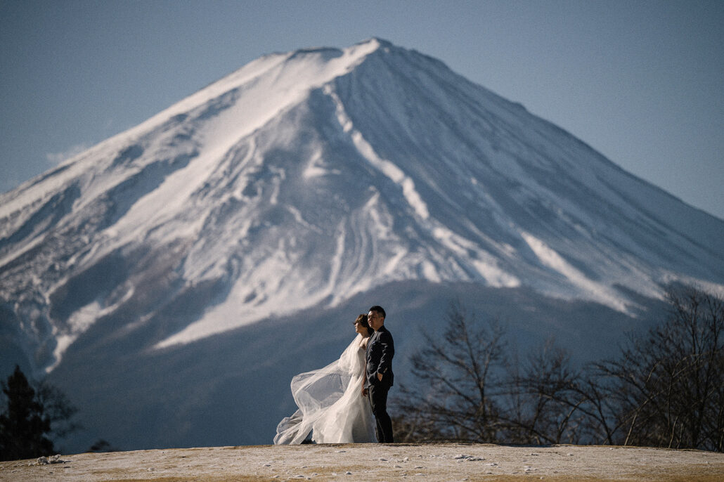 The bride's veil streams out as she stands with the groom on a bare hilltop, Mt Fuji close and white with winter snow behind them.