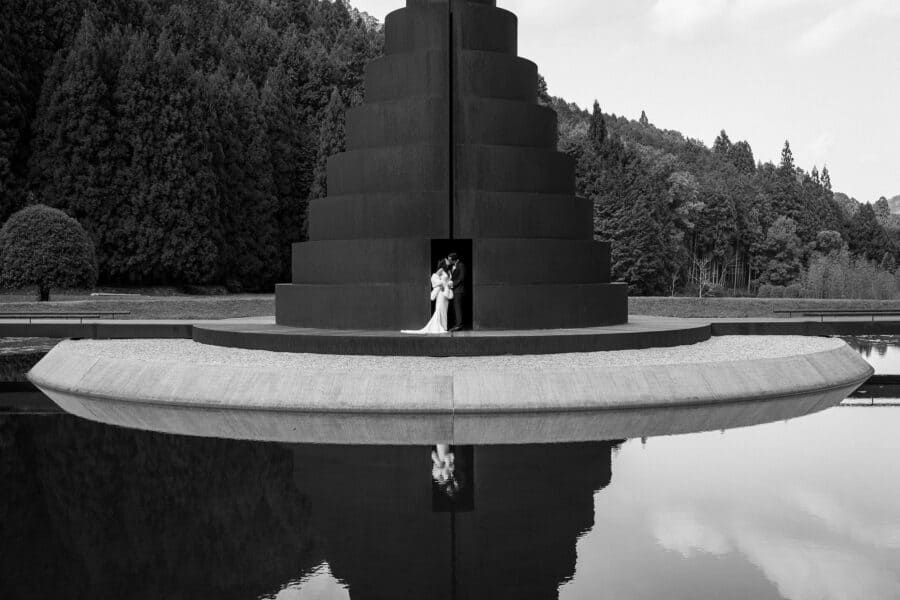 The couple stand in the doorway of a stepped steel tower mirrored in a still pond at Murou Art Forest, an elopement in Japan in black and white.