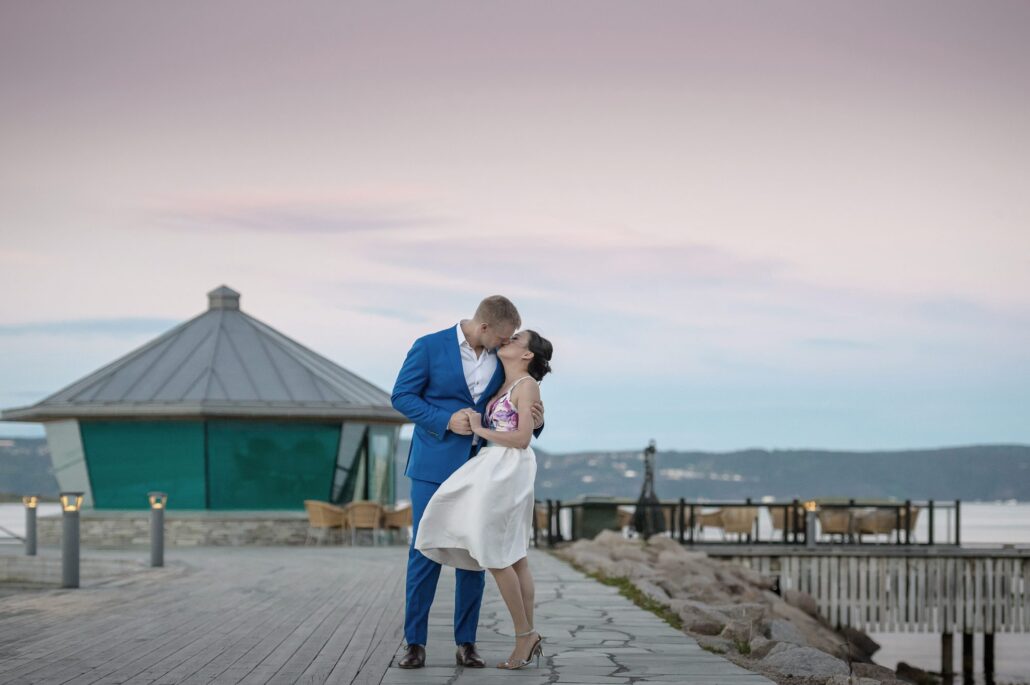 The bride and groom kiss on a waterside promenade in Norway, the sky pale pink behind them.