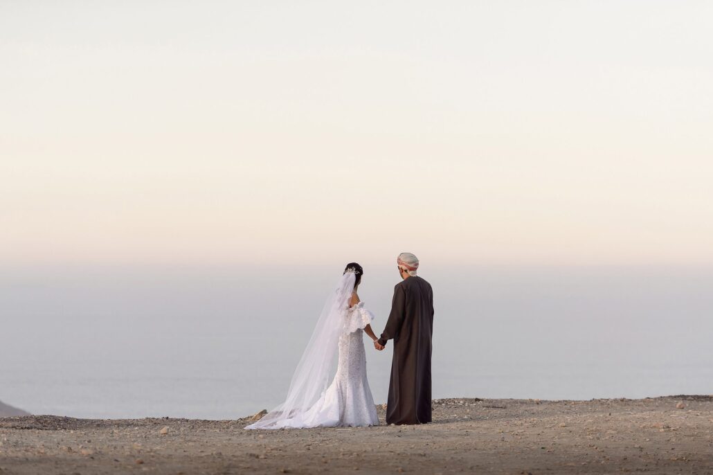 The bride and groom stand hand in hand on a clifftop above the sea at dawn, the groom in traditional Omani dress, at a destination wedding in Oman.