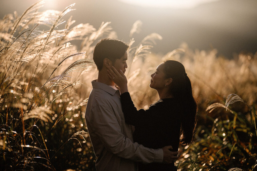 Close together in tall glowing grass, she holds his face in her hand as the autumn sun lights the plumes around them.