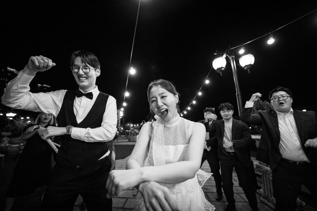 In black and white, the groom flexes an arm and the bride laughs as their friends dance beneath string lights.