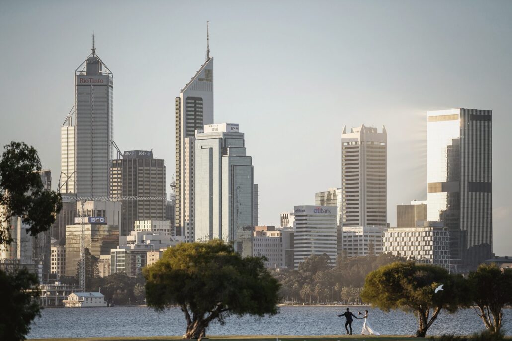 The couple stand small on the riverbank beneath the towers of the Perth city skyline.