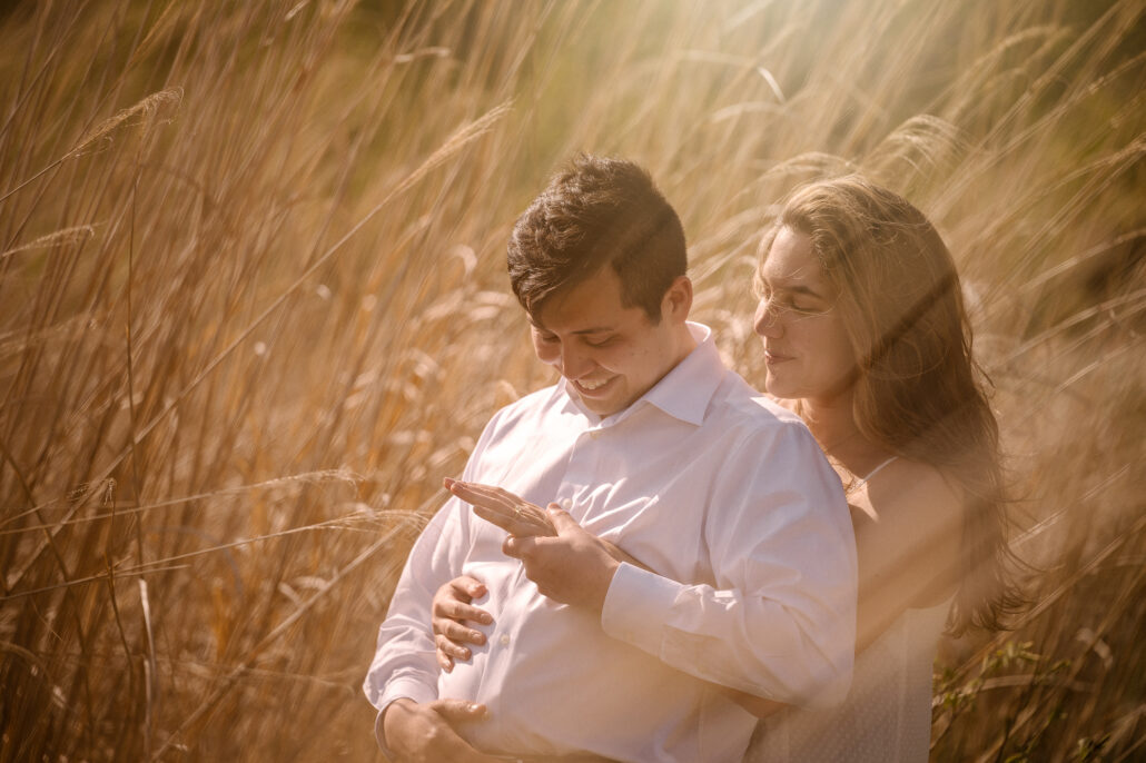 Holding him from behind, she watches as he smiles down at the engagement ring on her hand, tall gold grass glowing around them.