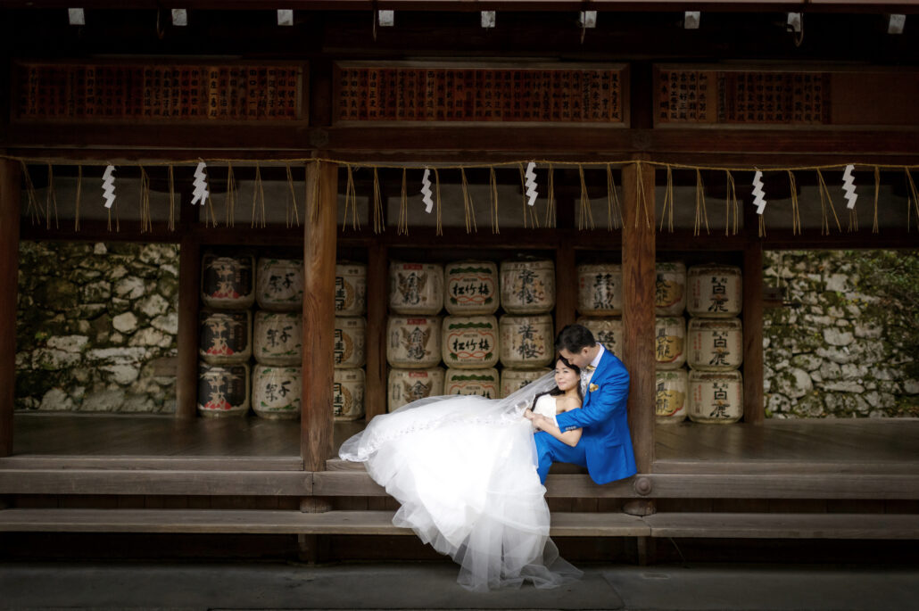 A couple sit together on the wooden veranda of a quiet Kyoto shrine, away from the crowds, a wall of sake barrels and paper streamers behind them.