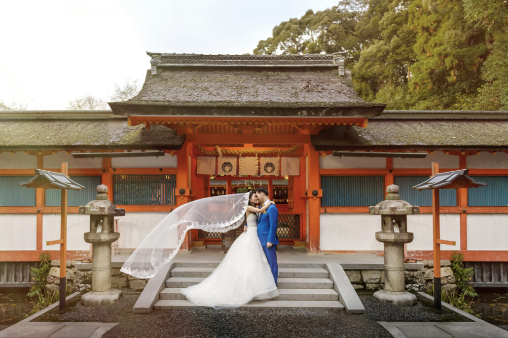 Wedding photos at a quiet Kyoto shrine: the couple embrace on the steps of a vermilion gate between stone lanterns, her veil lifting in the wind.