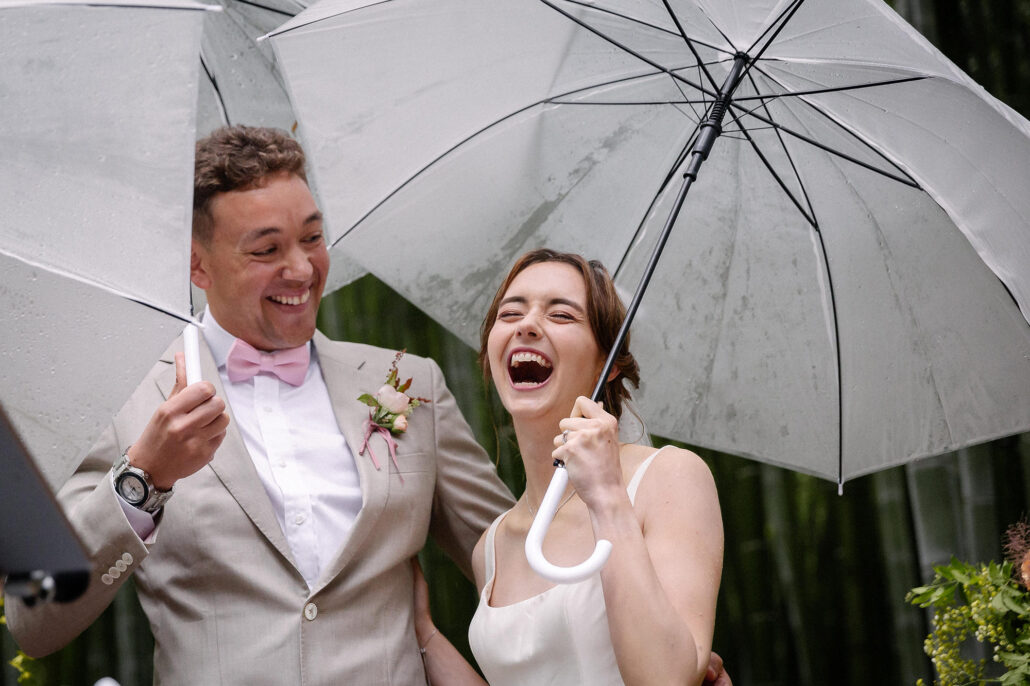 The bride and groom laugh together under clear umbrellas in the rain on their wedding day in a bamboo forest in Japan.