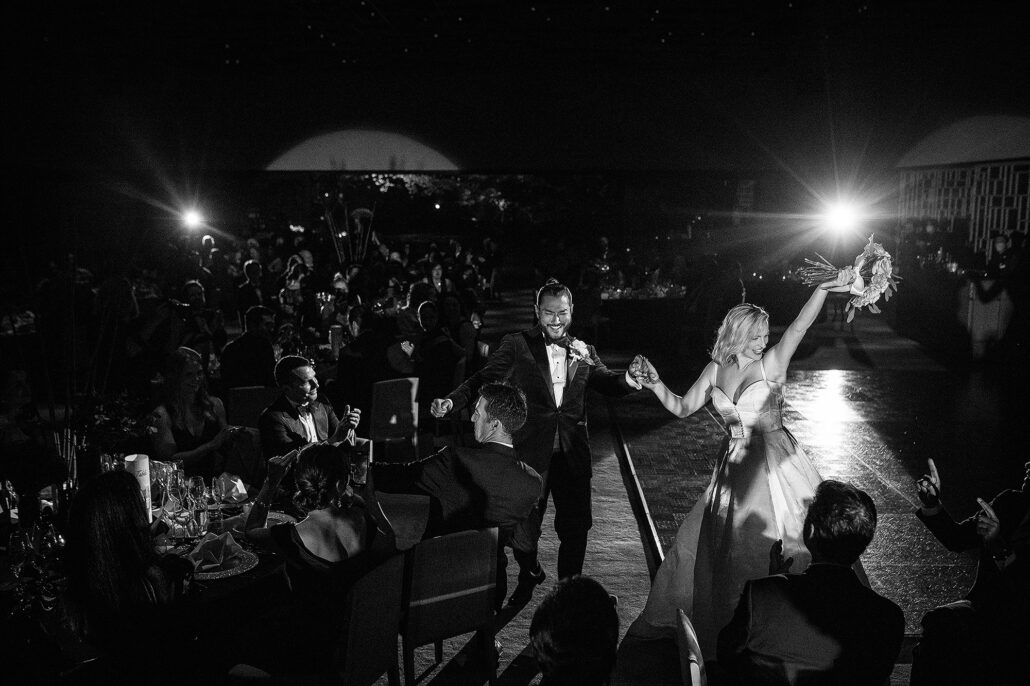 In black and white, the bride raises her bouquet as she and the groom dance into their reception between the tables.
