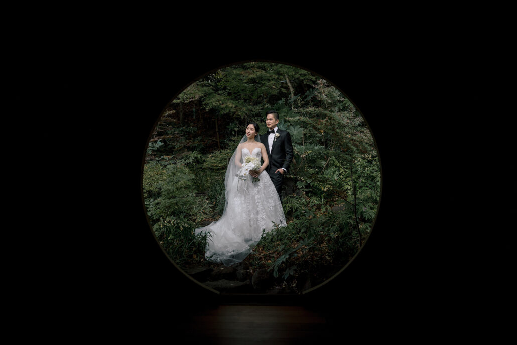 The bride and groom stand in the garden, framed by a round window in a dark wall.