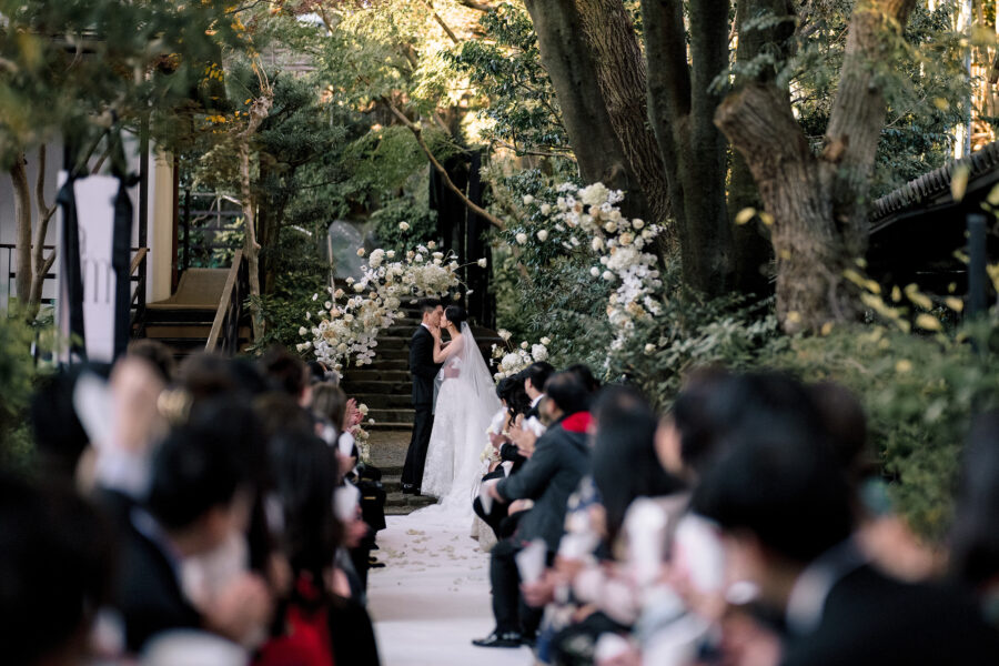 The couple kiss beneath a white floral arch at the top of the aisle as guests watch their outdoor ryokan wedding ceremony in Japan.