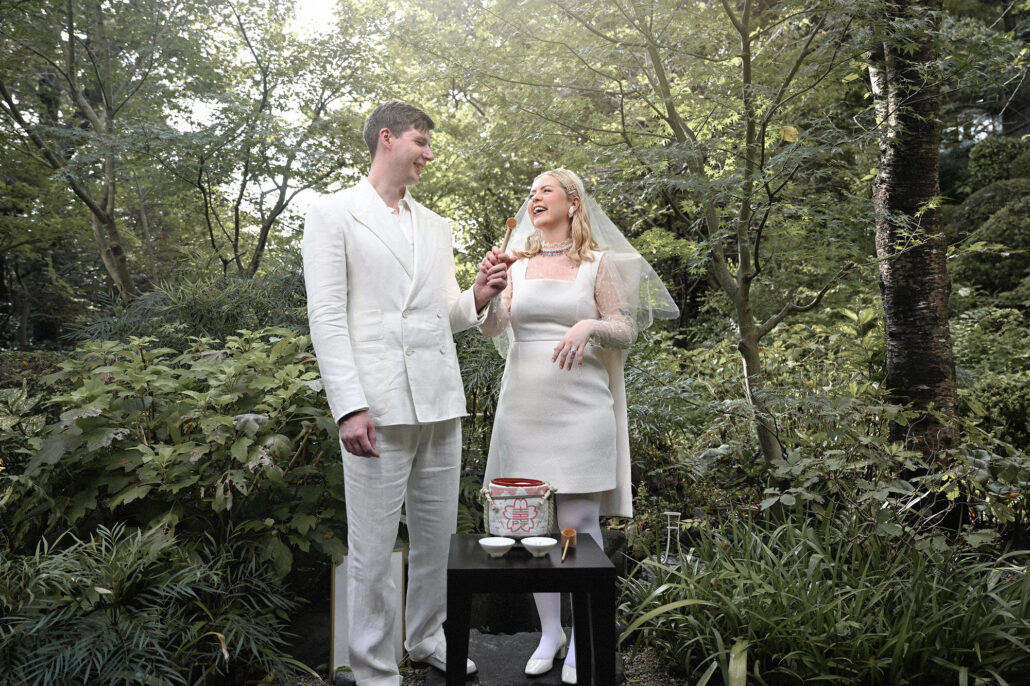 The bride laughs as she lifts a wooden sake ladle beside the groom, sake cups set on a small table in the elopement garden.