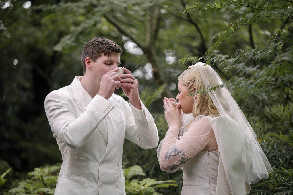 The bride and groom sip sake from small white cups under maple leaves, part of their elopement ceremony in Japan.