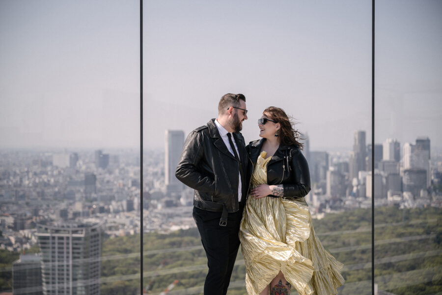 The bride in a gold pleated dress and the groom in a leather jacket laugh against the glass at Shibuya Sky, the Tokyo skyline behind them.