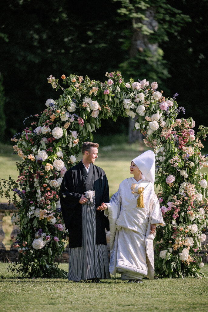 The bride in a white shiromuku and the groom in black montsuki walk out from a flower arch on a lawn at their wedding in France.