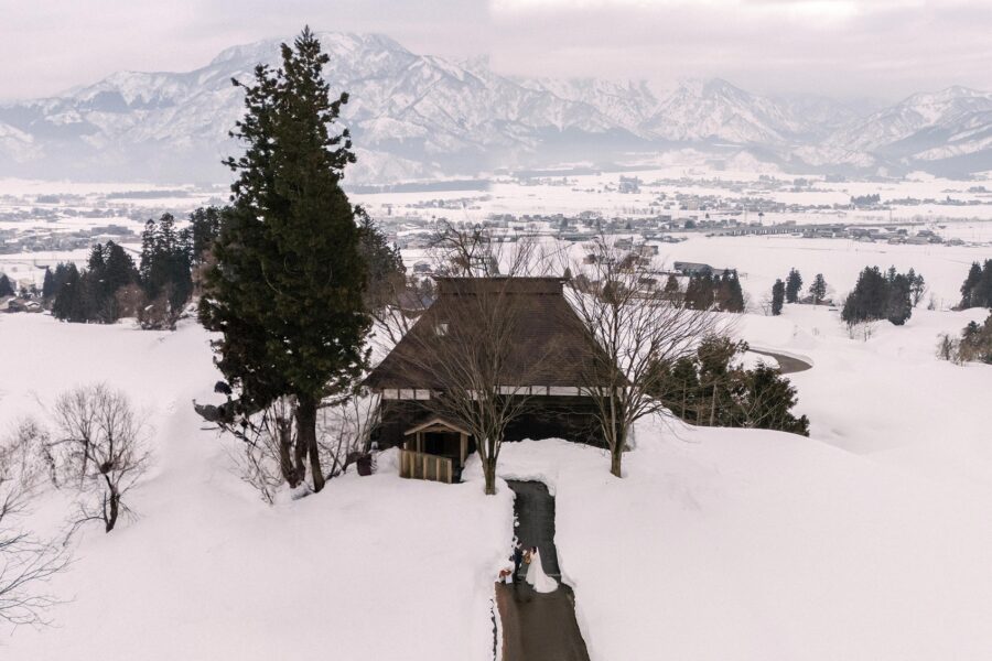 A drone view of the couple marrying on a path between two towering walls of snow below a traditional house, a snow elopement in Japan.