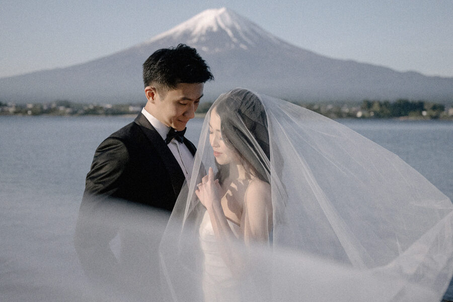 The couple stand at Lake Kawaguchi with her veil blowing across the frame and snow-capped Mt Fuji behind, spring pre-wedding photos.
