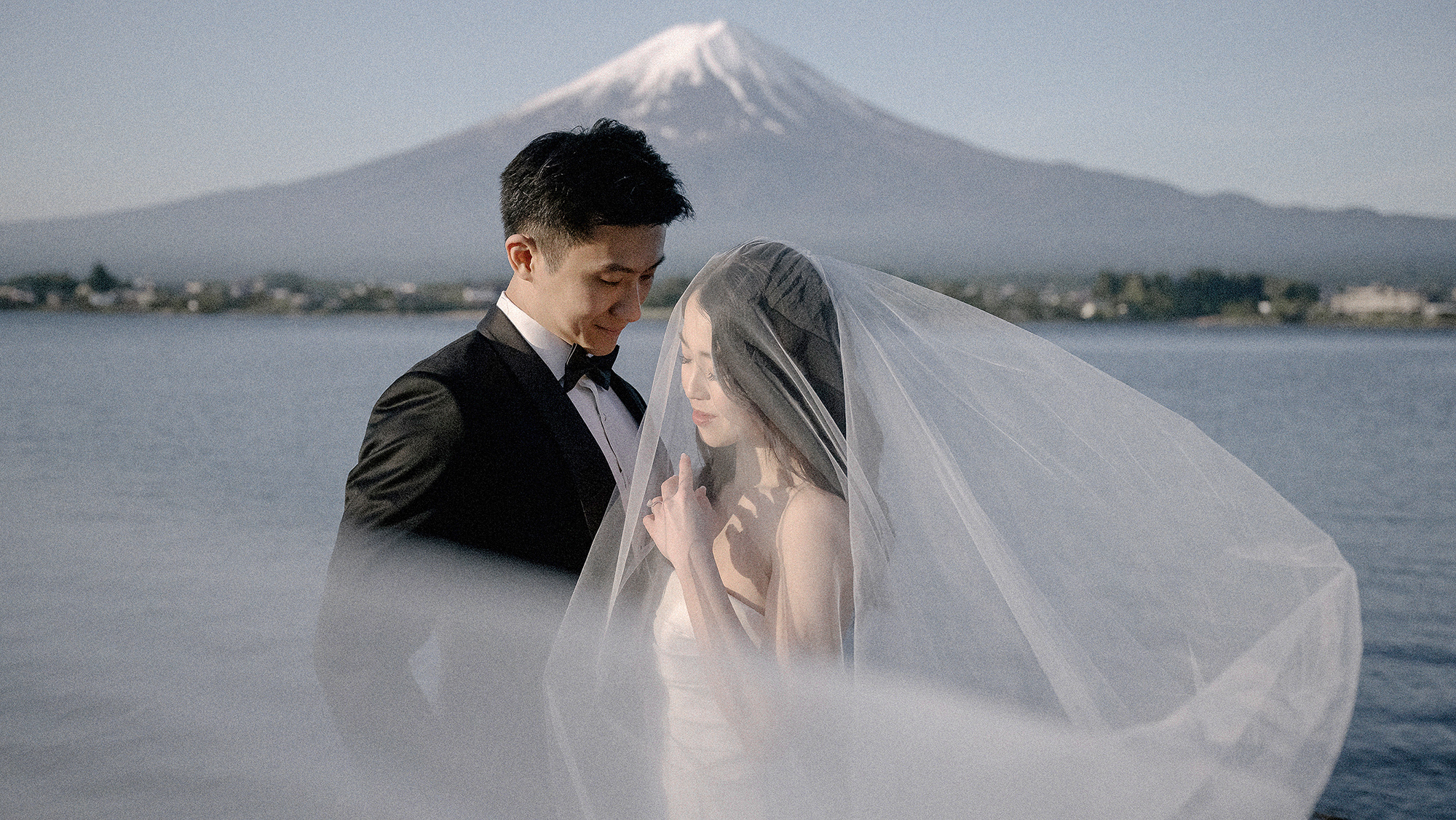 The couple stand at Lake Kawaguchi with her veil blowing across the frame and snow-capped Mt Fuji behind, spring pre-wedding photos.