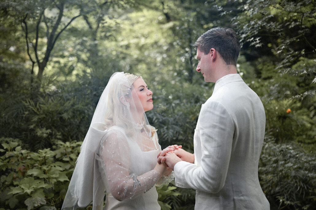 The couple face each other and hold hands during their summer elopement ceremony, the garden green and soft behind them.