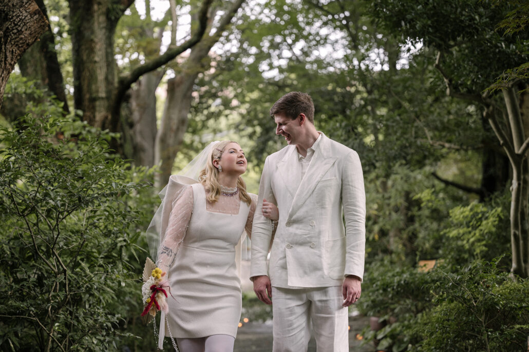 The bride and groom walk arm in arm through the old garden at Jinya, her handmade bouquet in hand, on their summer elopement in Japan.