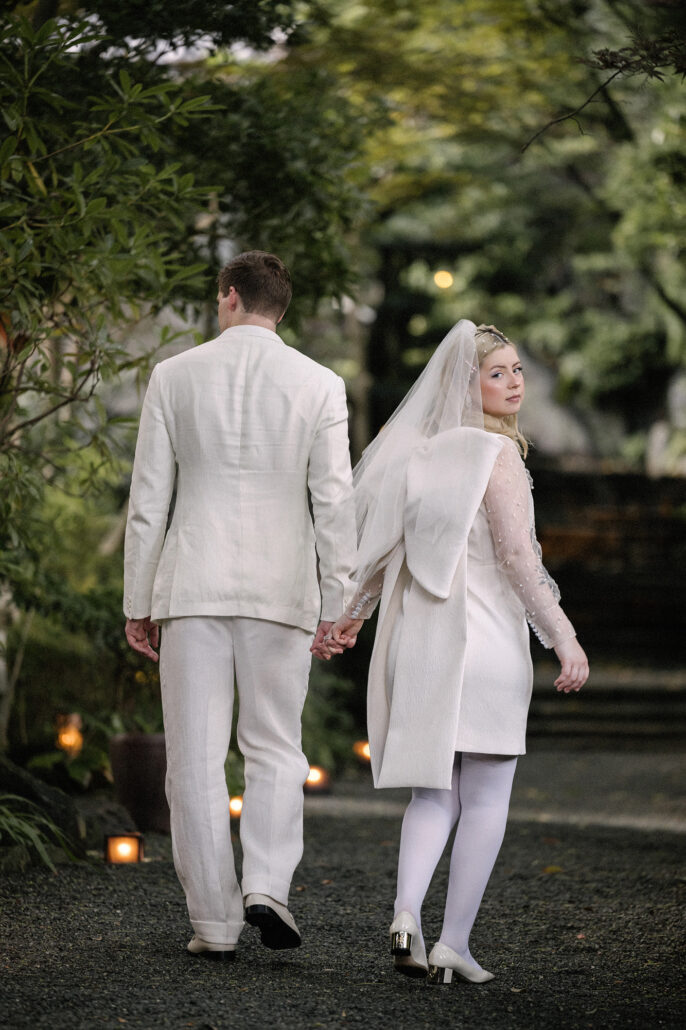 The couple walk away hand in hand down a garden path as the bride looks back, the end of their summer elopement in Japan.