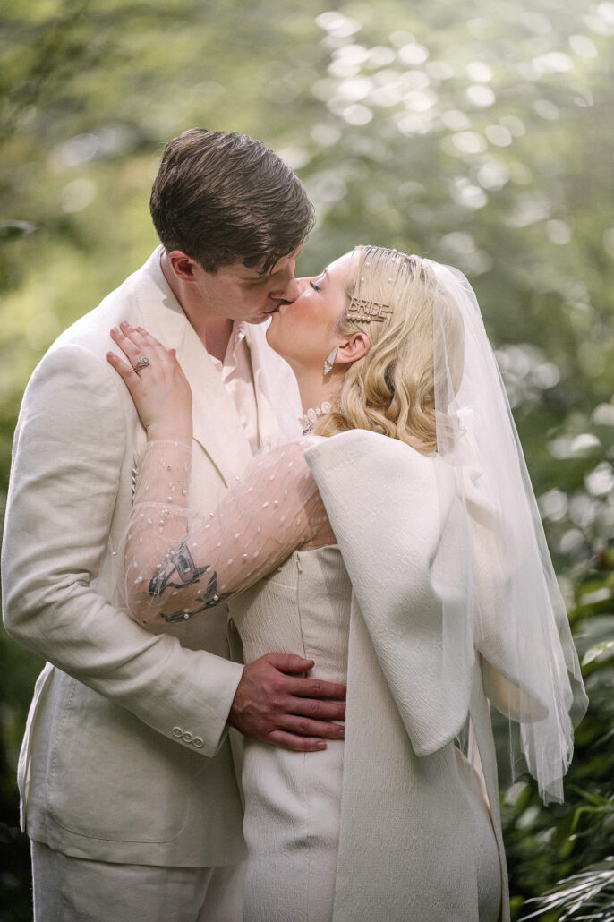 The bride and groom kiss among the leaves, her hand on his cheek, during their summer elopement in a Japanese garden.