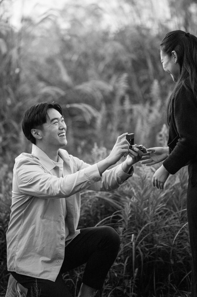 In black and white, he kneels in tall grass and holds out an open ring box as she reaches for it, smiling, by Lake Kawaguchi.